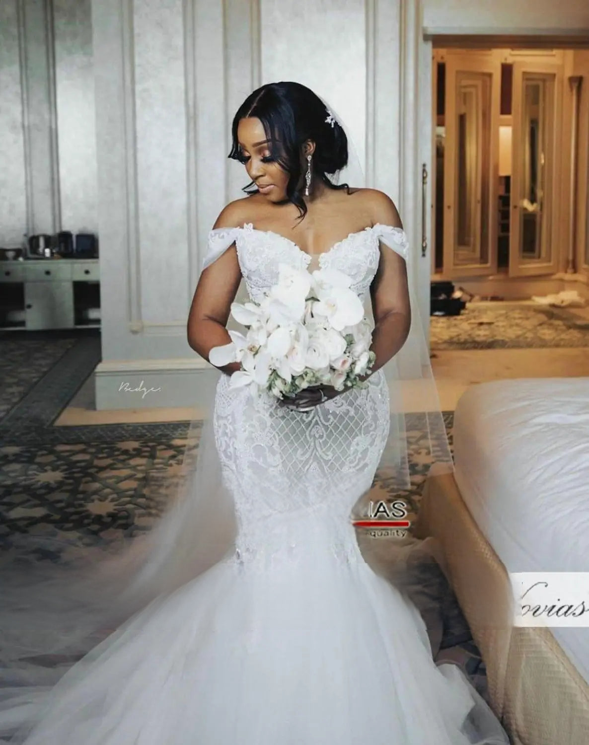 Beautiful African bride looking down at her white floral bouquet while wearing a fitted mermaid wedding dress with elegant lace detailing and sheer sleeves.