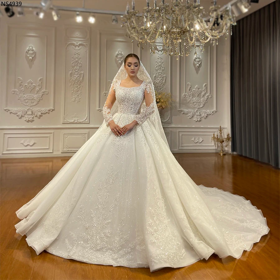 Full-length view of an elegant A-line wedding gown with a square neckline, sheer long sleeves, and intricate pearl embroidery throughout the bodice and skirt.