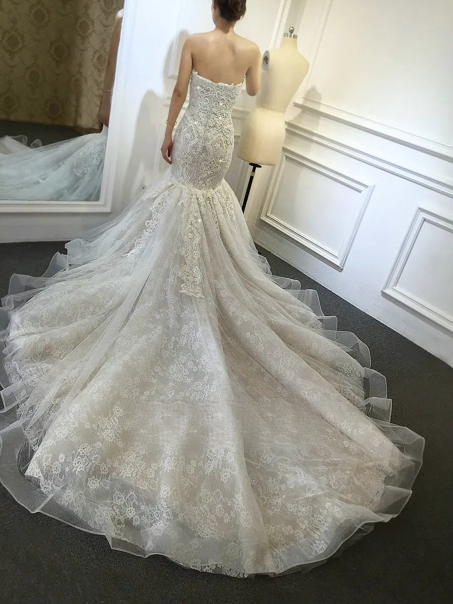 Rear view of a bride modeling an elegant champagne mermaid wedding dress, highlighting the sweeping, multi-layered lace train and intricate back beadwork, with her reflection visible in a mirror.