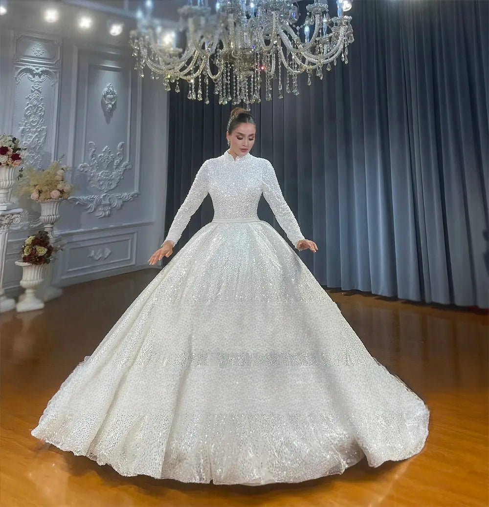 Grand full-length shot of a modest white ball gown wedding dress for Muslim brides, showing the voluminous sparkling skirt and long sleeves under a crystal chandelier.