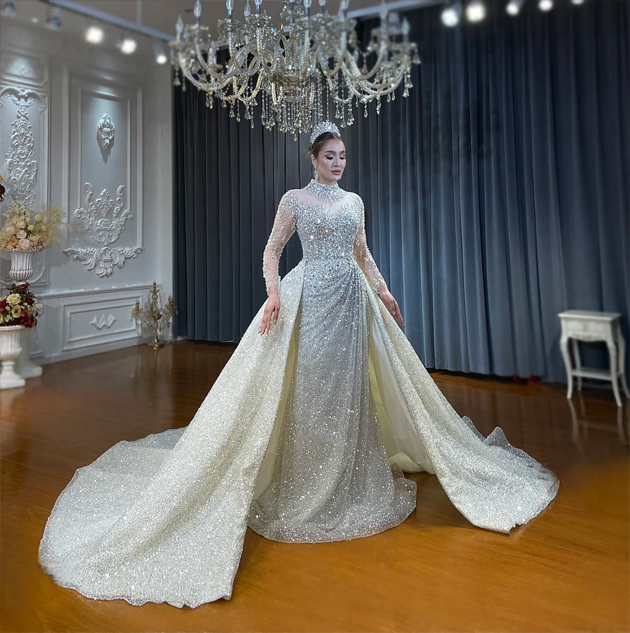 Full-length view of a model in the high-neck, long-sleeved crystal wedding dress with the overskirt, posing in a luxury bridal boutique under a chandelier.