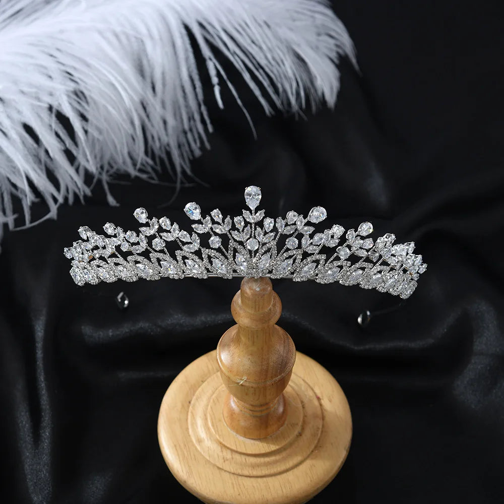 A luxury crystal bridal tiara featuring intricate leaf-like patterns, displayed on a wooden stand against a black background with white feathers.