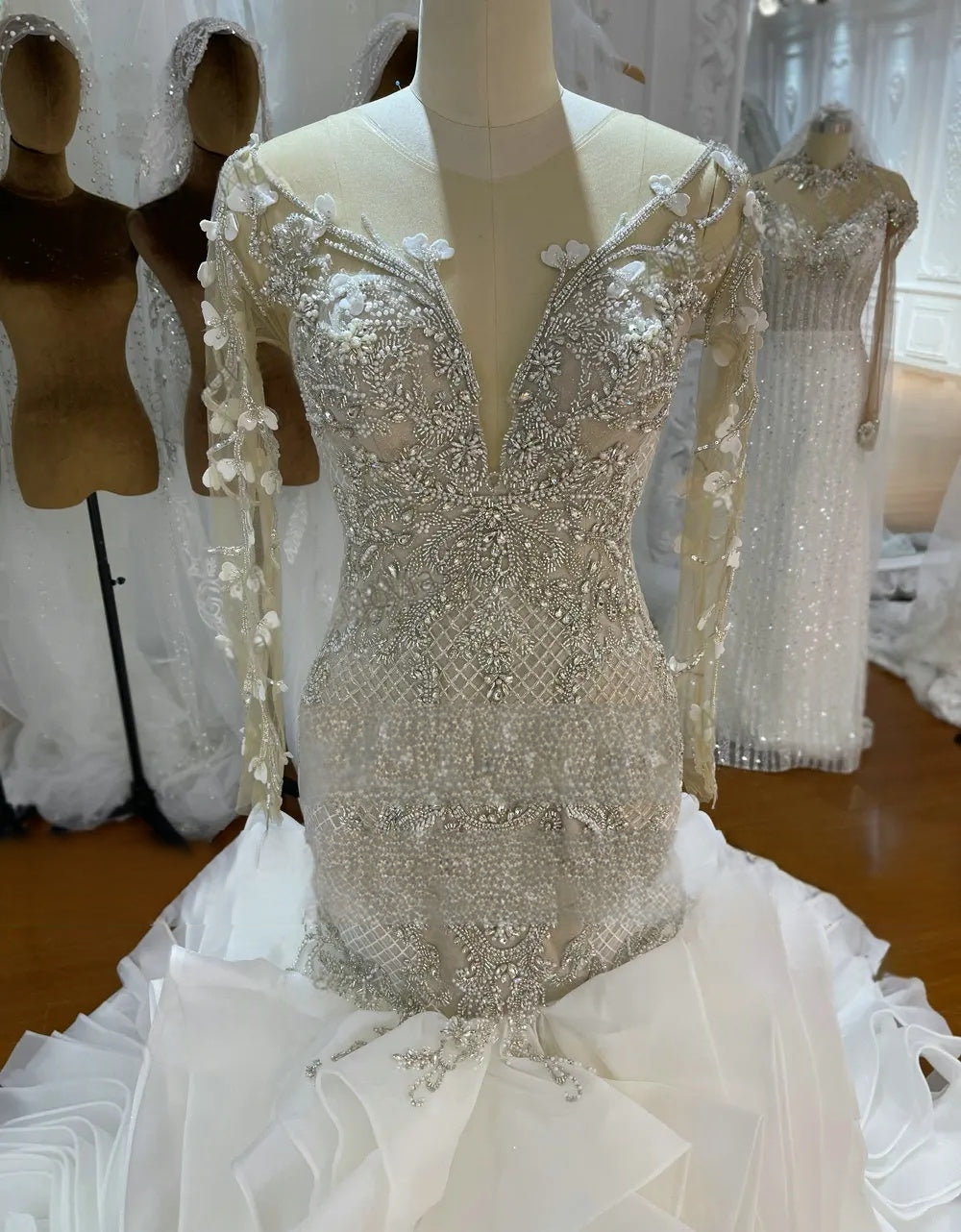 Close-up of a mermaid wedding dress bodice showcasing complex crystal beading and delicate 3D floral embroidery on sheer long sleeves.