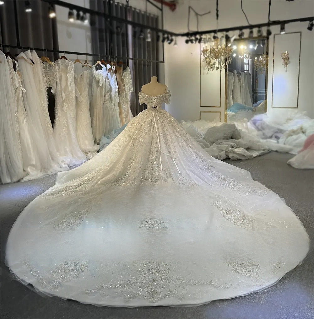Rear view of the elegant beaded ball gown on a mannequin, displaying the lace-up corset back, off-the-shoulder straps, and a massive shimmering cathedral train.