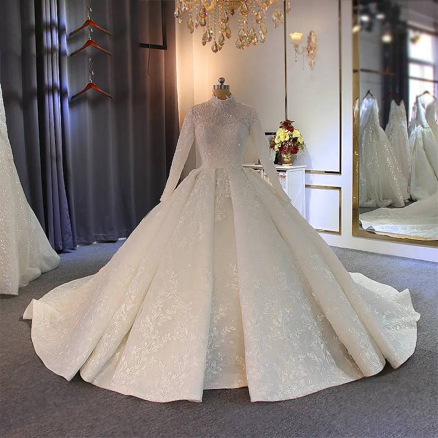 Mannequin display of a white modest wedding dress with long sleeves and a high neck, showcasing the dense floral lace patterns and full lining of the elegant ball gown.
