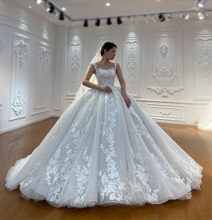 Romantic off-the-shoulder off-white wedding dress with a flowing silhouette and vertical floral lace patterns on a glitter tulle skirt.