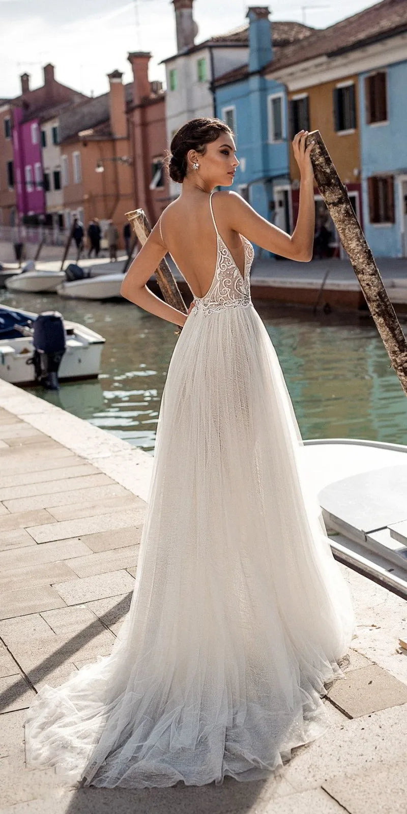 A back view of a bride posing by the water in a backless, pearl-detailed boho tulle wedding dress with a high side slit.