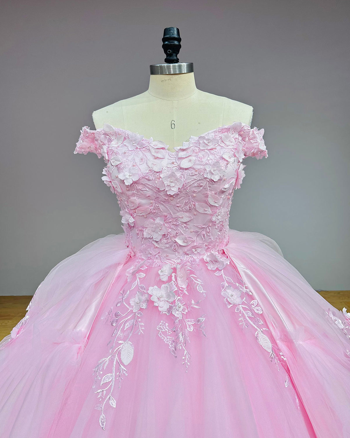 Showroom display of a blush pink off-the-shoulder wedding gown highlighted by intricate 3D flower details trailing down a voluminous tulle skirt.