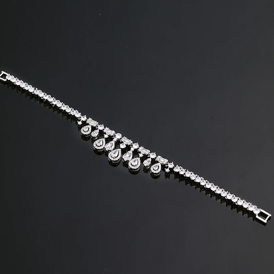 Diagonal flat lay view of a delicate silver cubic zirconia bridal fringe bracelet on a dark reflective surface.