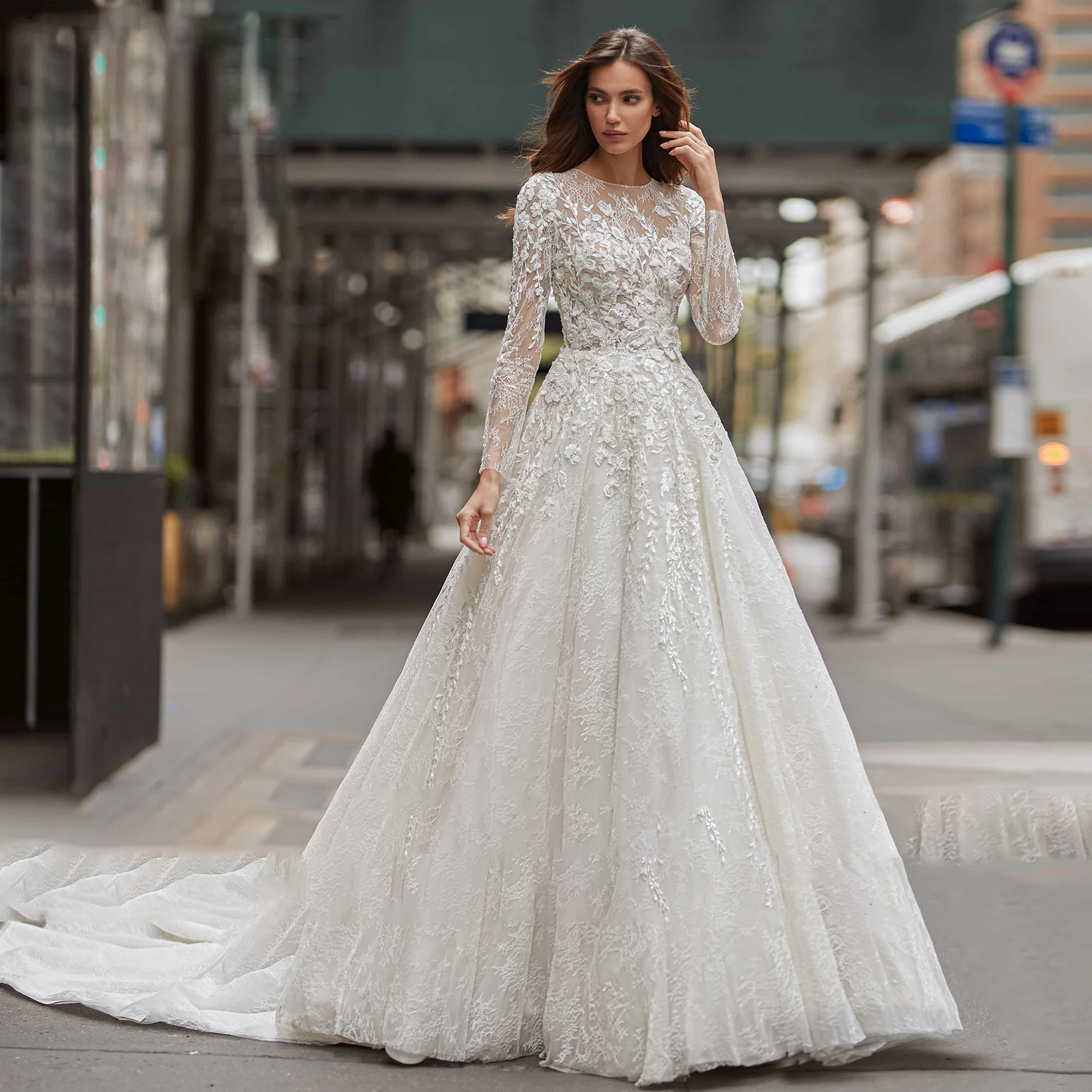 An elegant, full-length shot of a bride in an A-line long-sleeve wedding gown with sequinned beading and a brush train, posed outdoors in the city.