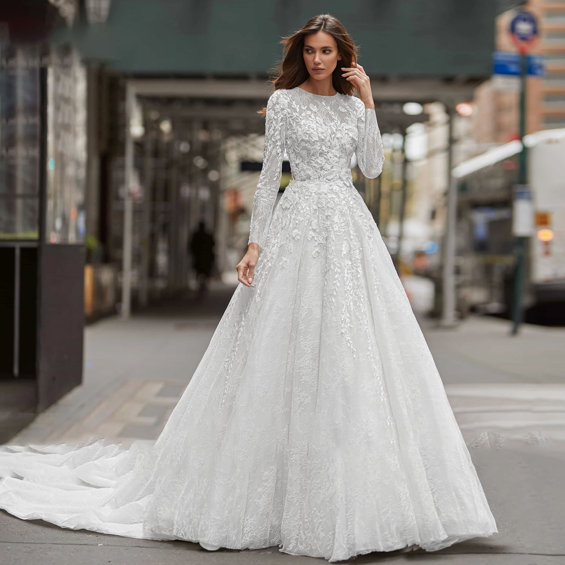 A full-length view of a bride wearing a long-sleeve A-line wedding gown, featuring a scoop neckline, sequinned illusion beading, and an elegant brush train on a city street.