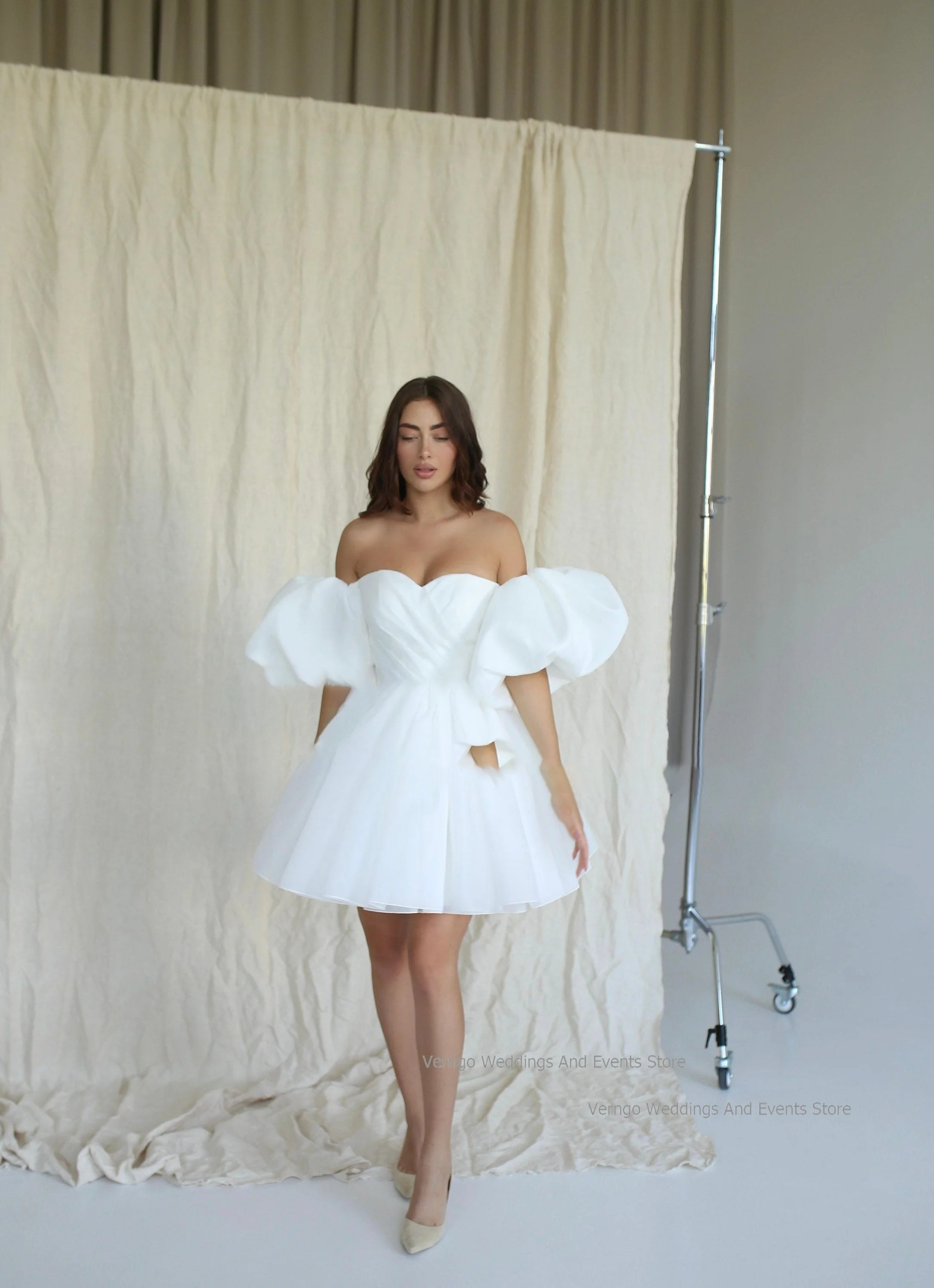 Full-length front profile of a modern short organza bridal gown with detachable puffy sleeves, styled as a chic mini reception dress against a studio backdrop.
