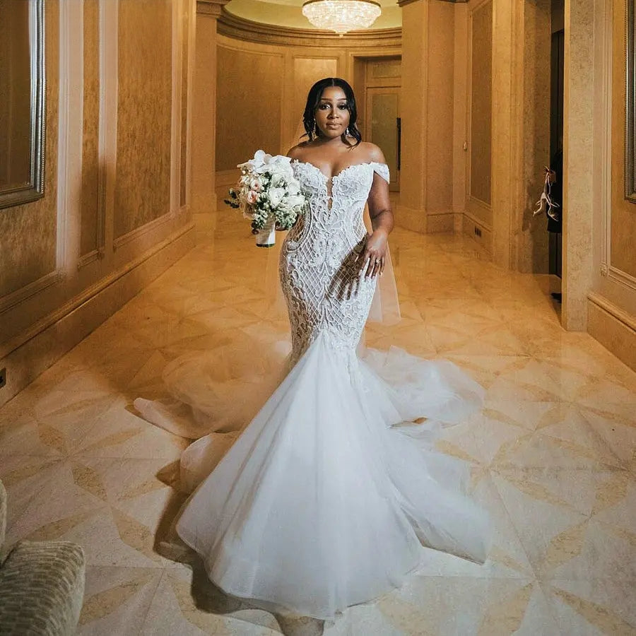 A stunning African bride wearing an off-shoulder mermaid wedding dress with elegant lace detailing and a voluminous tulle train standing in a grand hallway holding a white bouquet.