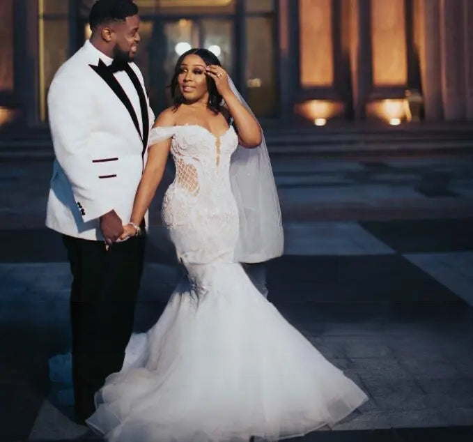 A romantic close-up of an African bride in a lace off-shoulder mermaid gown and her groom in a white tuxedo sharing a moment during their wedding.