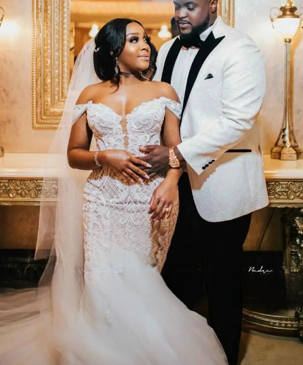 Elegant portrait of a bride wearing a custom African off-shoulder mermaid wedding dress with exquisite lace detailing posing with her groom.