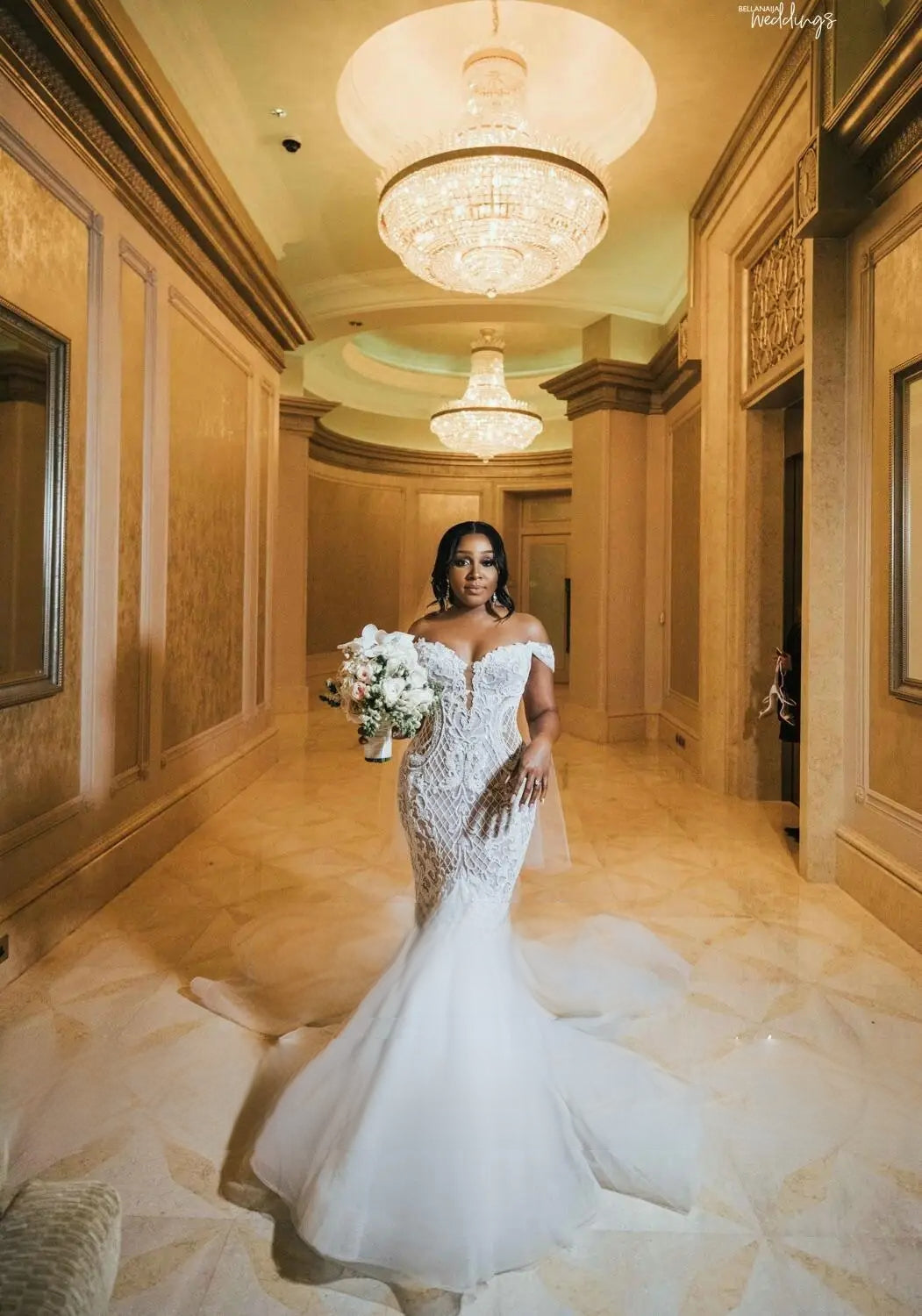 Full-body view of a bride in an African-inspired off-shoulder mermaid bridal gown with a grand train standing under luxurious chandeliers.