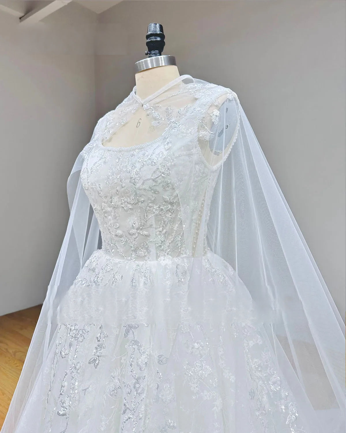 Detailed side profile of a bridal gown bodice on a mannequin, highlighting the layered tulle cape and heavy beadwork along the neckline and shoulders.