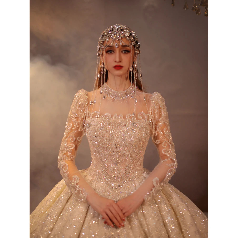Close-up of a 2025 luxury ball gown wedding dress with a square neckline and long illusion sleeves, featuring heavy crystal beading and a matching royal jeweled headpiece.