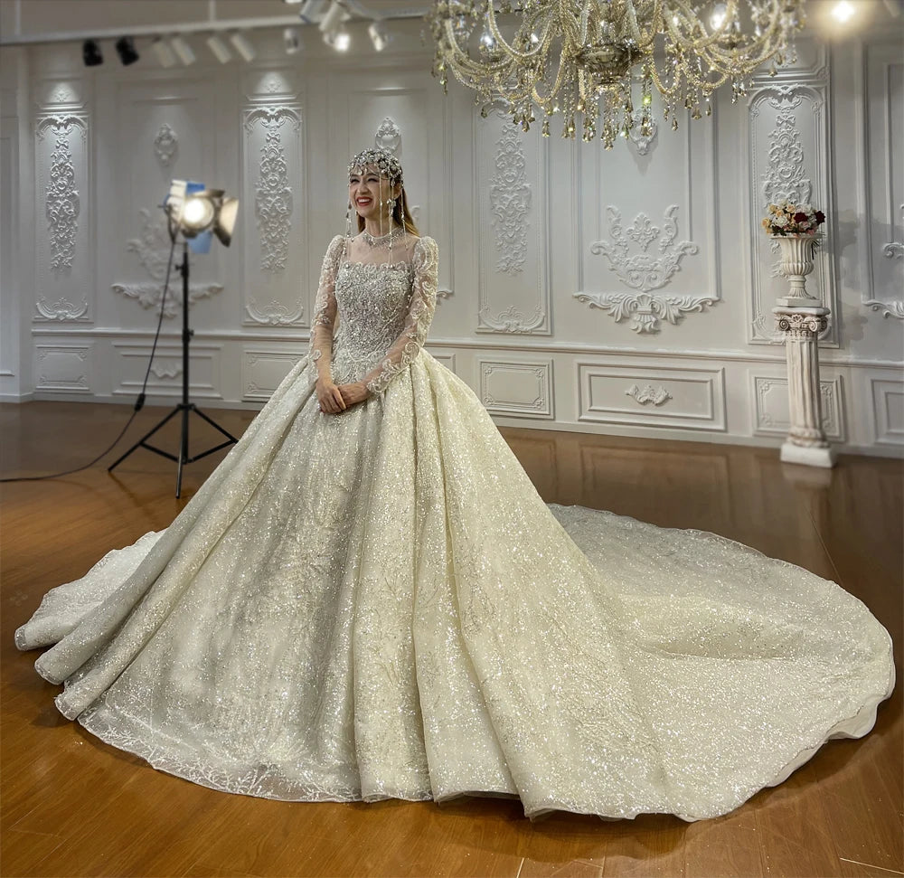 Elegant bride in a silver-toned shimmering ball gown wedding dress with a majestic full skirt and grand train, posed in a luxury bridal showroom under a chandelier.
