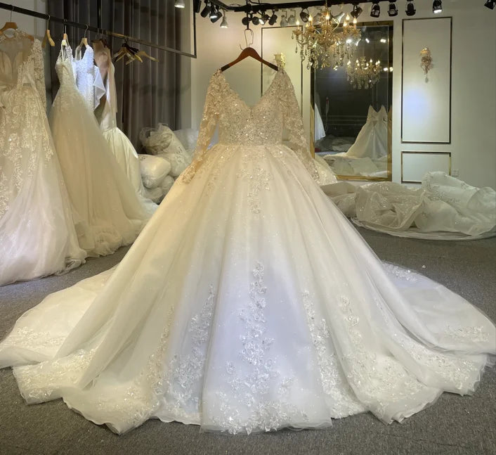 Stunning front perspective of a luxury long sleeve bridal gown with shimmering tulle and floral lace appliques, showcased by a model posing under a grand crystal chandelier.