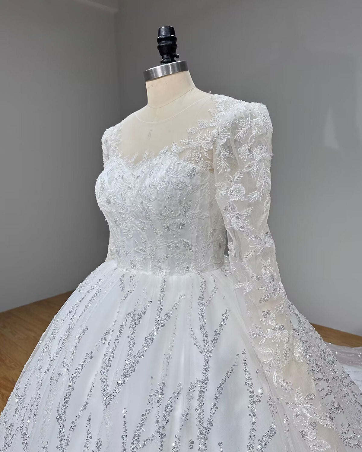 Three-quarter close-up of a luxury wedding gown bodice highlighting sheer long sleeves with floral lace appliqués and abstract sparkling sequin branches.