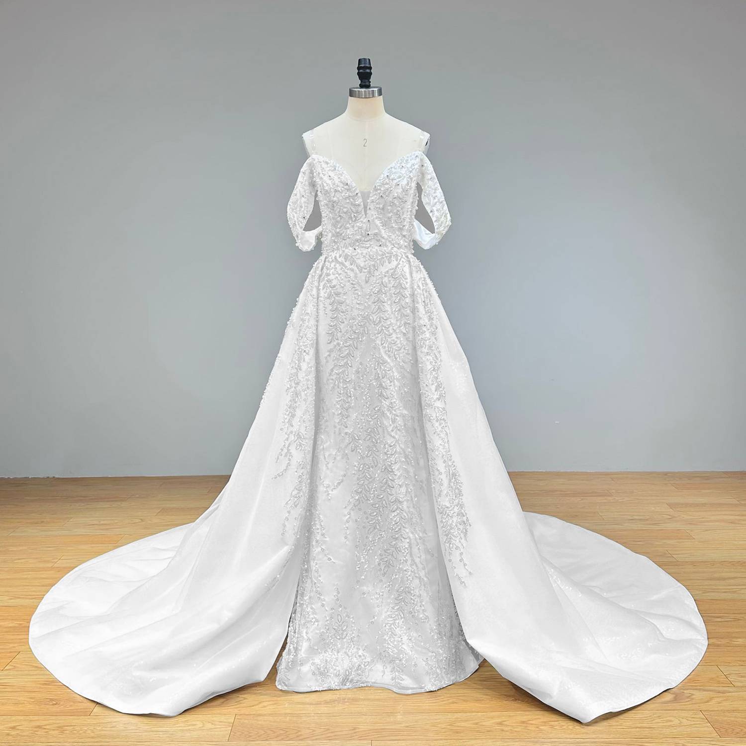 Wide front studio shot of an off-the-shoulder beaded lace mermaid wedding dress on a mannequin, showcasing the timeless bridal elegance and the grand scale of the detachable train.