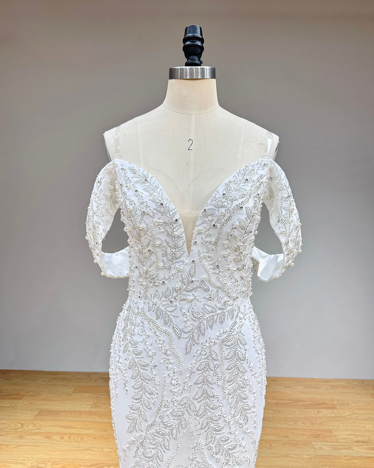 Close-up front bodice detail of a mermaid bridal gown on a mannequin, emphasizing the off-the-shoulder design, plunging neckline, and intricate beaded lace embroidery.