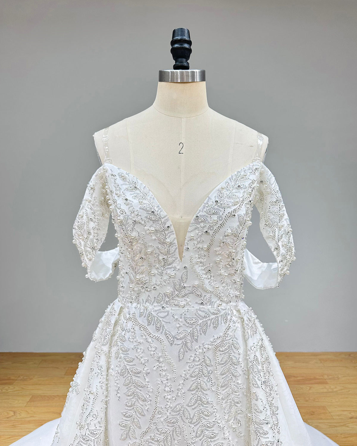 Close-up front bodice detail of a mermaid bridal gown on a mannequin, emphasizing the off-the-shoulder design, plunging neckline, and intricate beaded lace embroidery.