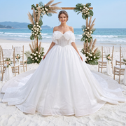 Glamorous bride wearing a luxury glitter tulle ball gown wedding dress with a pearl-beaded corset and an off-the-shoulder bow neckline, posing on a scenic beach wedding set.