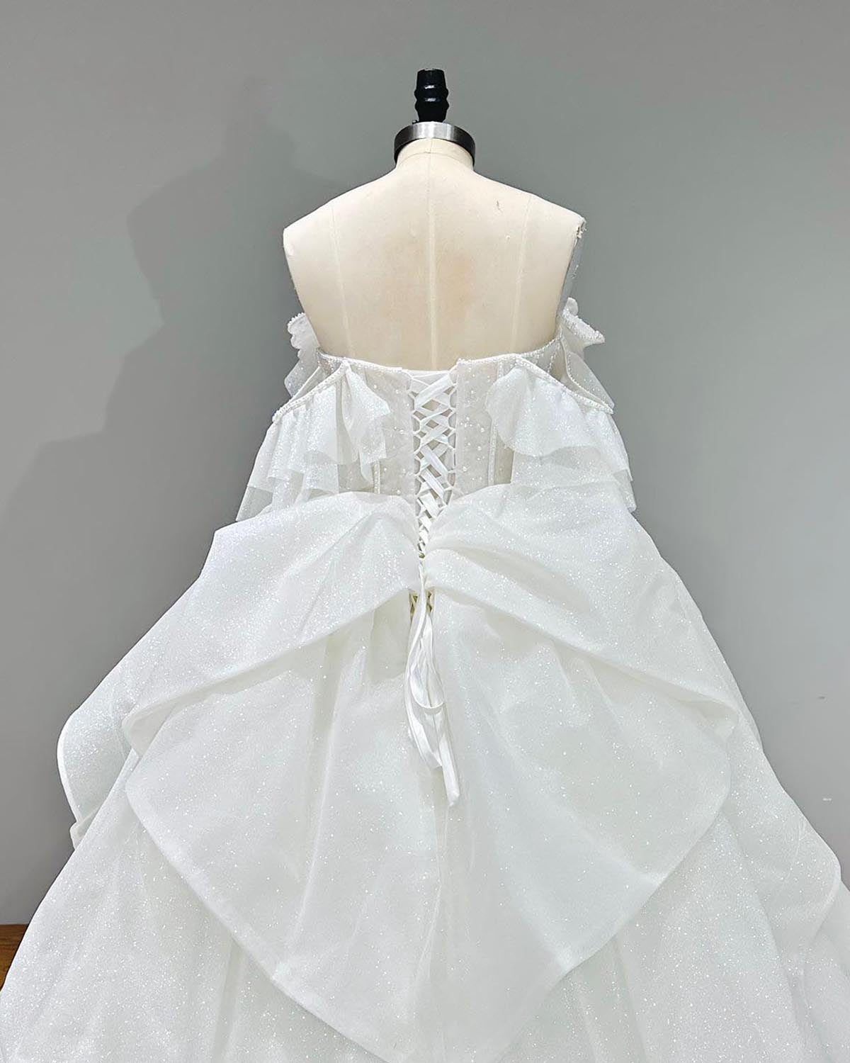 Detailed upper back view of a modest bridal gown on a dress form, showcasing the adjustable satin ribbon corset closure transitioning into a voluminous tiered skirt attachment.