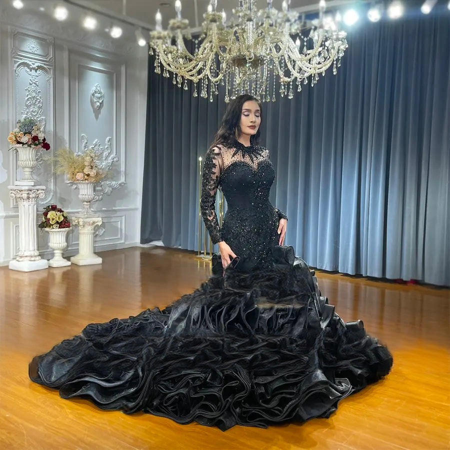 Side-profile view of a model wearing a stunning black gothic-style mermaid wedding dress, highlighting the beaded lace sleeves and the dramatic texture of the large, tiered ruffled skirt in a luxury showroom setting.