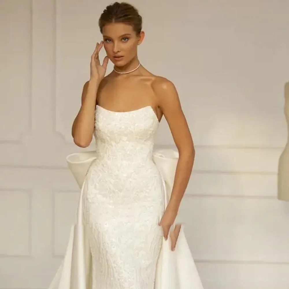 Close-up front view of a strapless white wedding gown bodice highlighting structured sequin detailing and a dramatic detachable satin overskirt bow back.