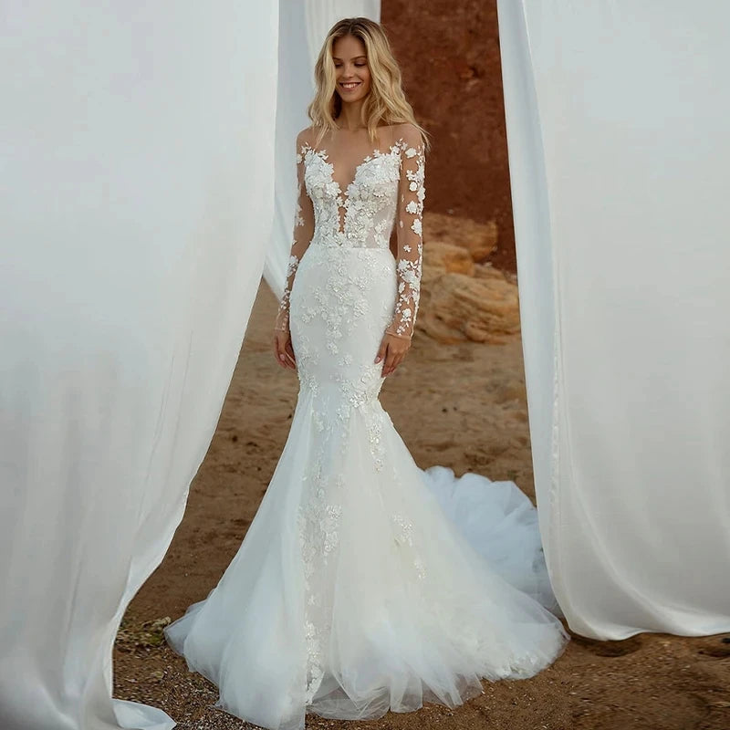 A bride modeling a bohemian-inspired mermaid wedding dress with intricate lace appliqués, a V-neckline, and sheer long sleeves on a sandy beach.
