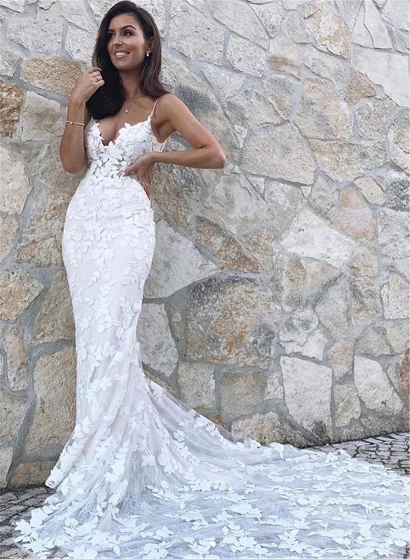 A bride posing against a stone wall, showcasing the tight-fit mermaid silhouette, lace appliqués, and deep neckline of this boho bridal gown.