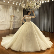 Full-length front view of a luxury long sleeve ball gown wedding dress featuring a shimmering, voluminous skirt and intricate silver beadwork, showcased under a grand crystal chandelier.