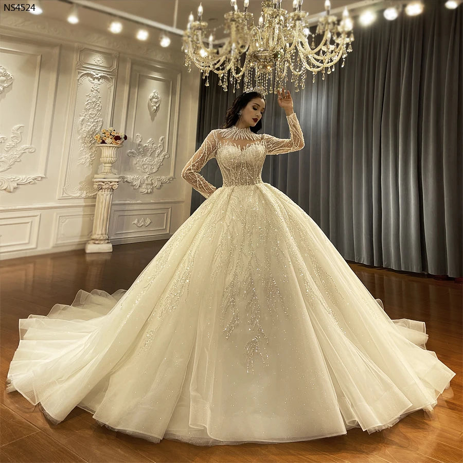 Full-length front view of a luxury long sleeve ball gown wedding dress featuring a shimmering, voluminous skirt and intricate silver beadwork, showcased under a grand crystal chandelier.