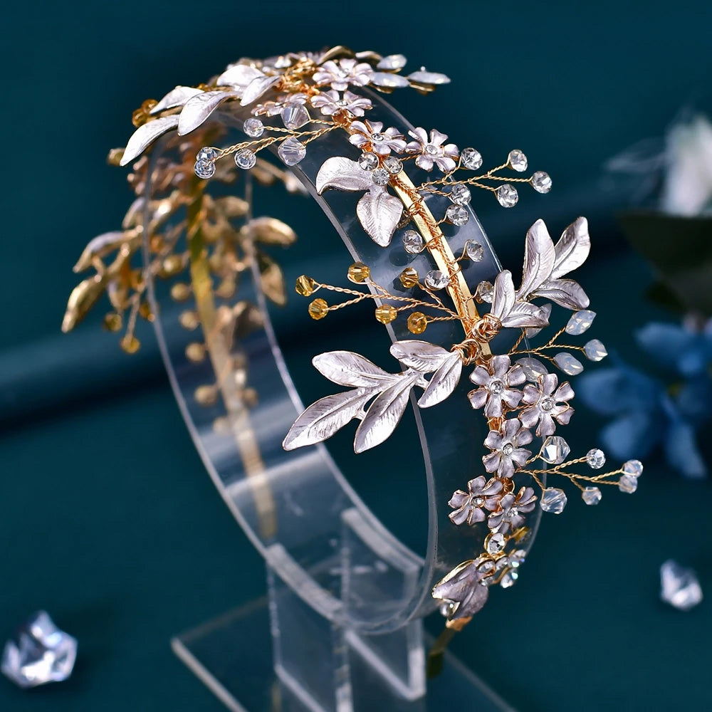 Side angle of a gold bridal flower headband with textured silver leaves and clear crystals displayed upright on a transparent acrylic headband stand.