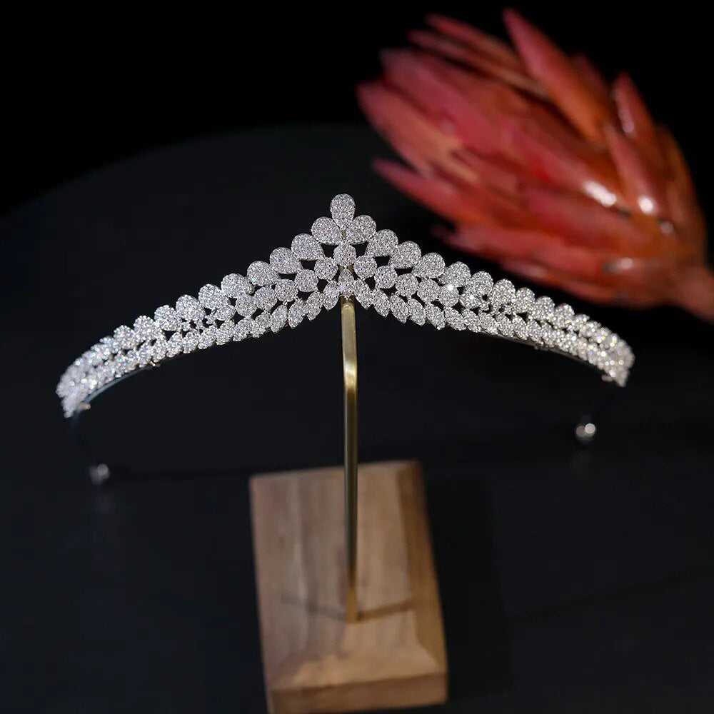 Front perspective of a luxurious zirconia wedding crown tiara showcased on a retail display stand, highlighted by a decorative reddish floral prop in the background.