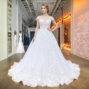 Full-length front view of a model wearing a white cap sleeve A-line wedding dress with intricate floral appliques and a shimmering skirt in a luxury bridal studio.