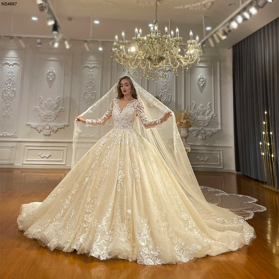 Full-length front view of an elegant model wearing a champagne full lace ball gown wedding dress with sheer long sleeves and an intricate V-neckline, holding a matching cathedral-length lace veil in a luxury bridal showroom.