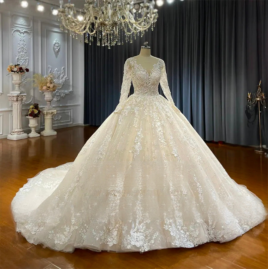 Front perspective of an exquisite champagne bridal gown on a mannequin, showcasing detailed floral lace appliques, long sheer sleeves, and a voluminous ball gown skirt under a crystal chandelier.
