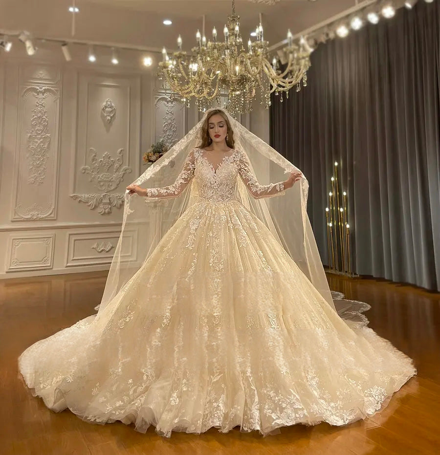 A radiant model showcasing a long sleeve lace ball gown wedding dress in champagne, highlighting the shimmering lace embroidery and the grand scale of the tulle skirt paired with a sheer bridal veil.