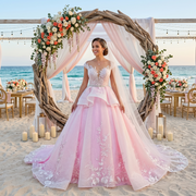 Smiling bride wearing a dramatic pink ball gown wedding dress with a white lace bodice and tiered peplum skirt, standing before a beachside floral wedding arch.
