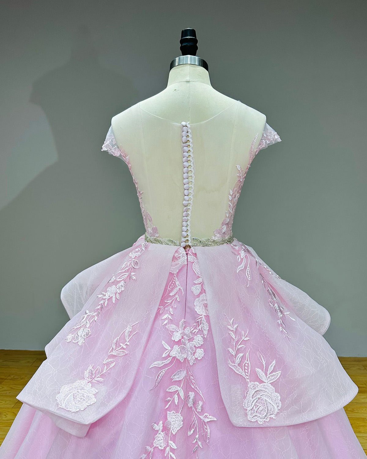 Close-up back view of the pink special event gown on a mannequin, highlighting the sheer illusion back with fabric-covered buttons and a crystal beaded waist belt.