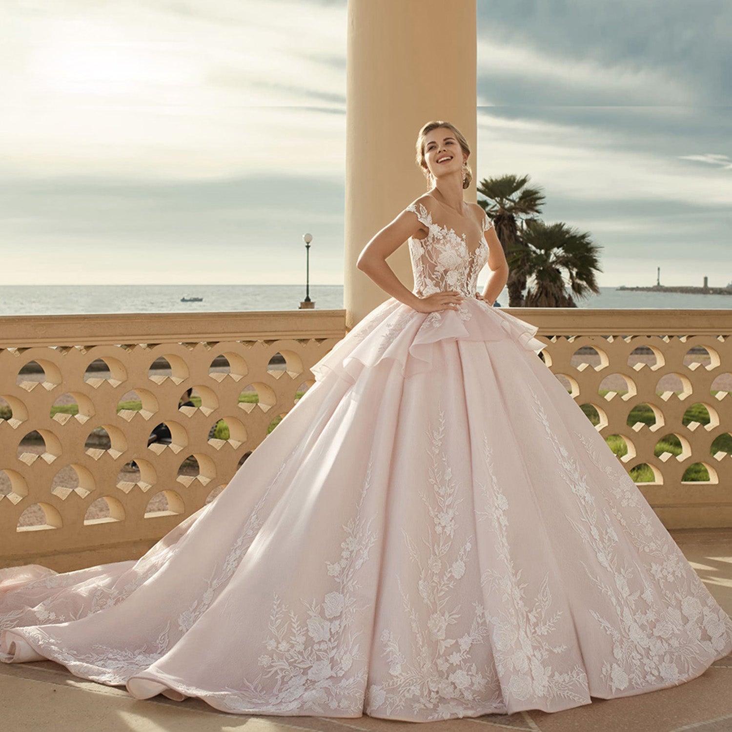 Editorial view of a smiling bride posing on a seaside terrace in a voluminous pastel blush wedding ball gown with delicate white floral embroidery and cap sleeves.
