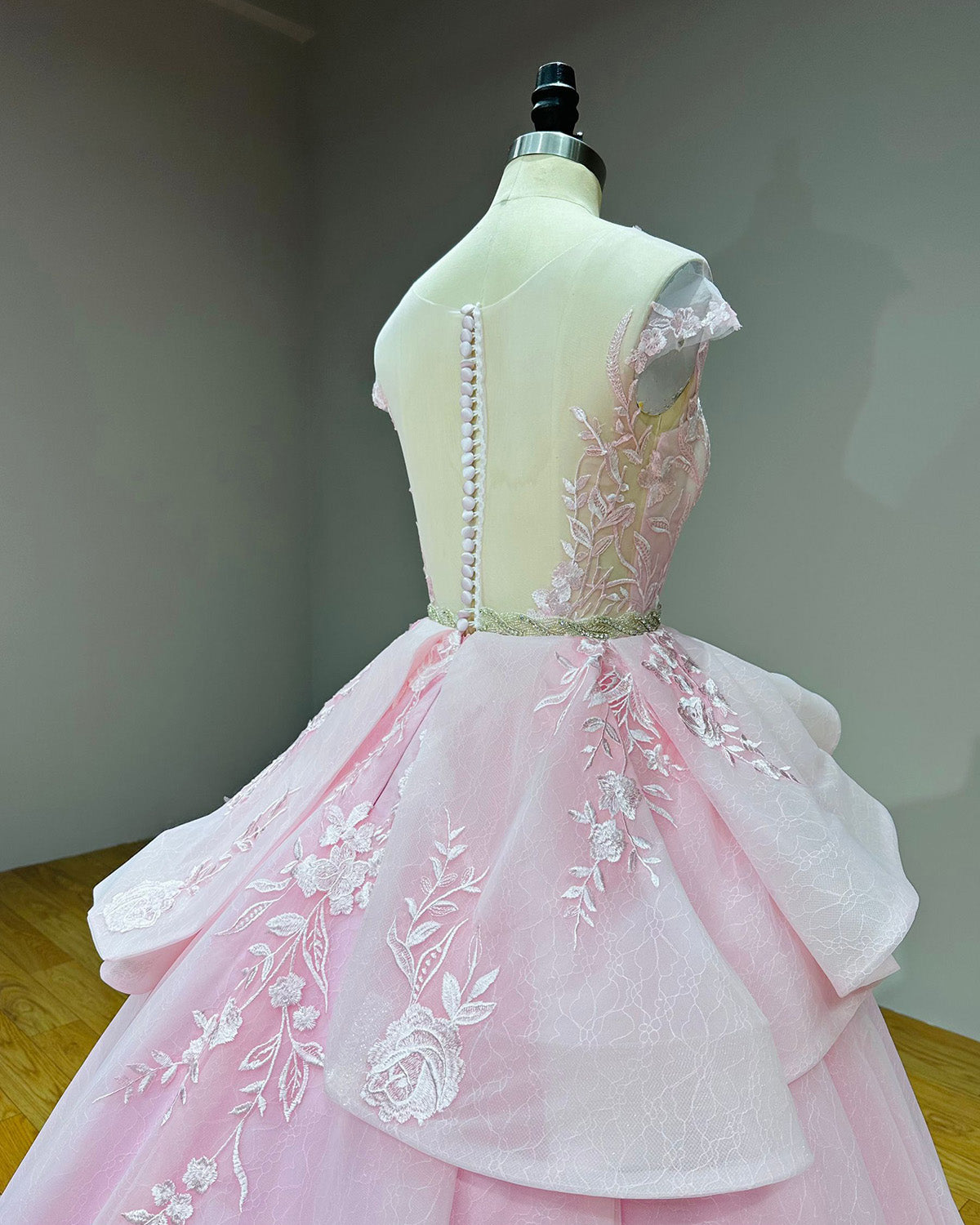 Three-quarter back angle detail of the blush pink bridal gown bodice on a mannequin, showing the sheer mesh back, button alignment, and cascading peplum folds.