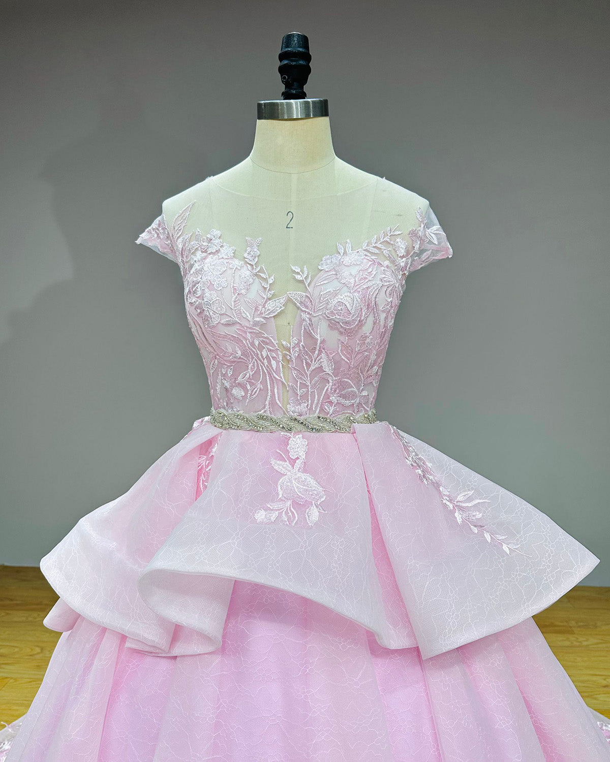 Close-up front view of the wedding dress bodice on a mannequin, highlighting the sheer illusion plunge neckline, white floral lace appliques, and a shimmering crystal belt.