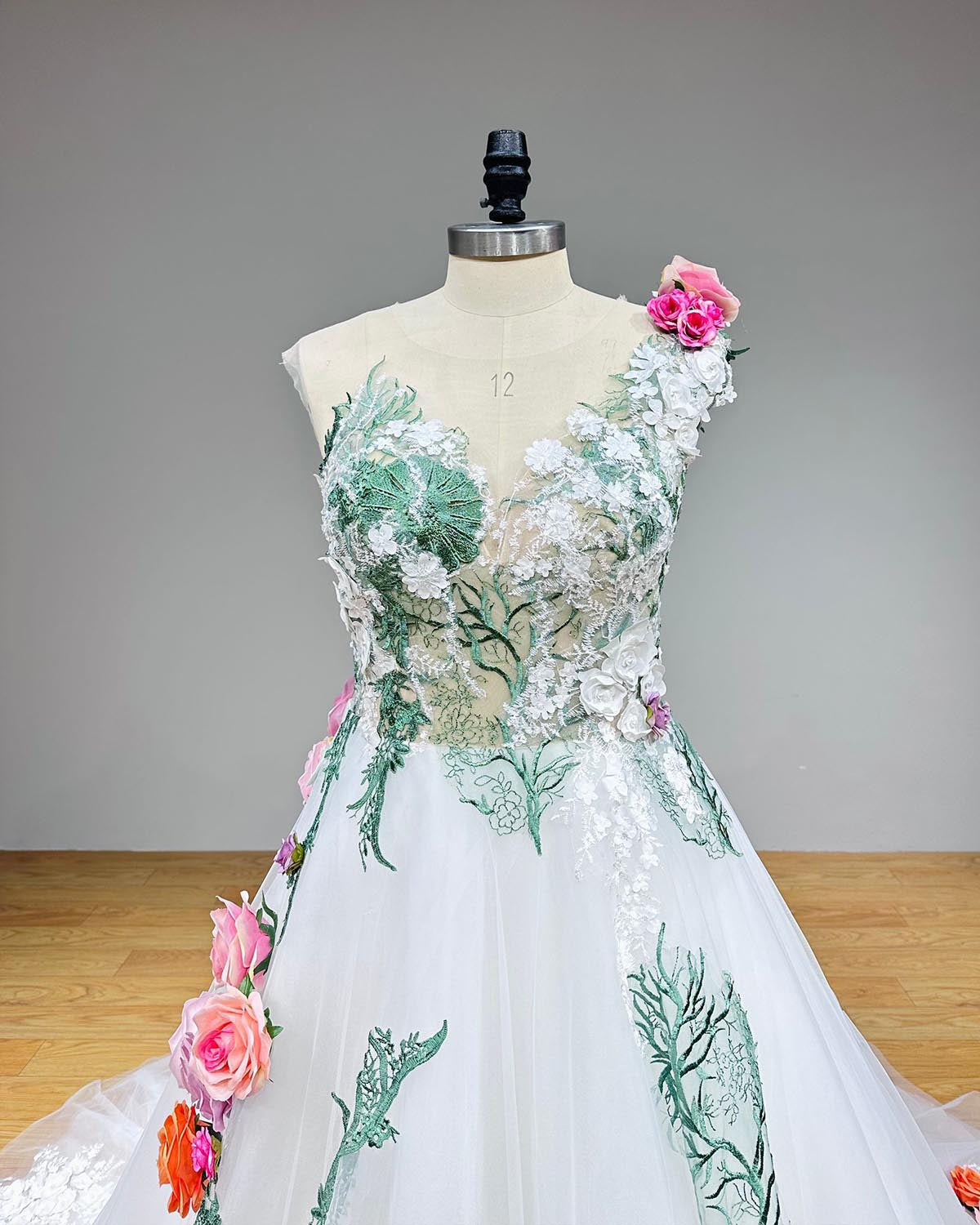 Close-up front bodice detail of a colorful tulle wedding dress on a mannequin, detailing the sheer illusion sweetheart neckline, green vine embroidery, and vibrant 3D flowers on the asymmetrical shoulder strap.