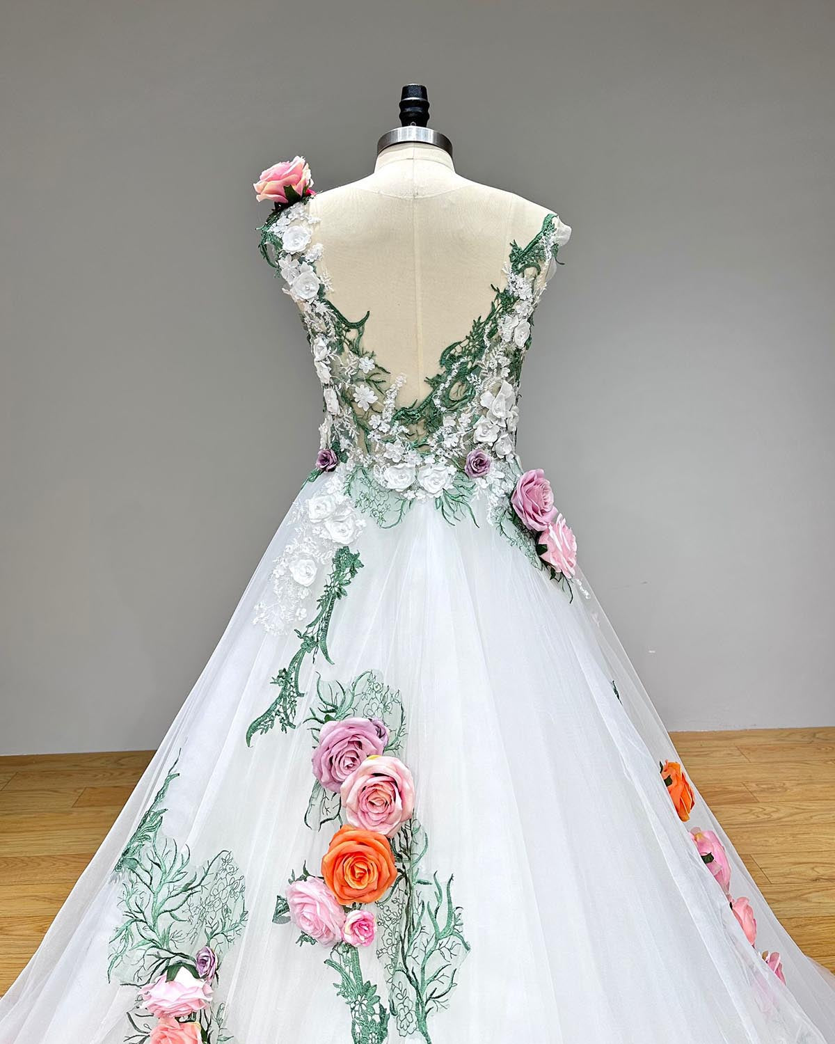 Close-up back bodice view of a colorful tulle ball gown wedding dress on a mannequin, displaying the elegant sheer open V-back design adorned with delicate green lace and trailing 3D flowers.