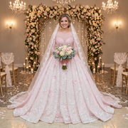Smiling bride wearing a pastel pink crystal beaded ball gown wedding dress with long sheer sleeves, standing before a floral altar holding a rose bouquet.
